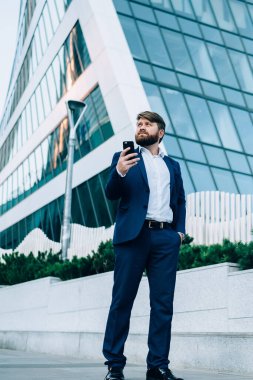 Zarif takım elbiseli, düşünceli, kendine güvenen bir yöneticinin caddede dikilirken ve akıllı telefonu şehir merkezindeki modern yüksek binalara karşı kullanırken gözlerini kaçırıp karar vermesinden.