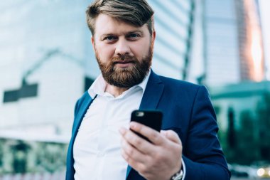 Resmi giyinmiş, kendine güvenen sakallı bir adam gün batımında bulanık gökdelenlere karşı sokakta akıllı telefonla iletişim kurarken kameraya bakıyor.