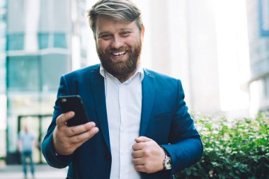 Mavi takım elbiseli içerik sakallı erkek yönetici, arka planda bulanık binalarla cep telefonuyla mesajlaşırken eğleniyor.