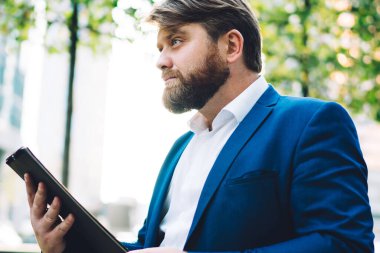 Kenarlarında ağaçlar ve ofis binalarıyla şehir merkezinde dikilirken elinde tablet olan düşünceli, kendine güvenen yönetici erkeğin yan görüntüsü.