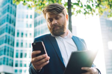 Şehir merkezindeki ofis binasının yanında elinde tabletle dururken cep telefonundan mesaj okuyan modern profesyonel erkek müdürden.