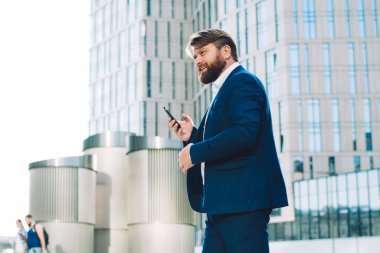 Şehir merkezinde caddeyi geçerken cep telefonu kullanan ve mesajları kontrol eden mavi takım elbiseli, meşgul yönetici sakallı adamın yan görüntüsü