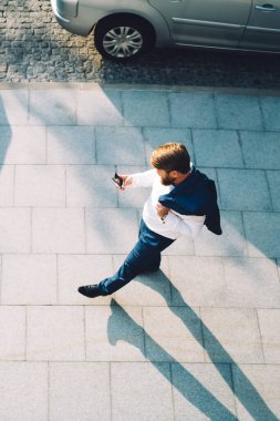Odaklanmış mavi takım elbiseli, omuzlarının üzerinde ceketini taşıyan, güneşli şehirde asfalt yolda yürürken akıllı telefonuyla dolaşan işadamlarının tepesinde gölge bırakıyor.