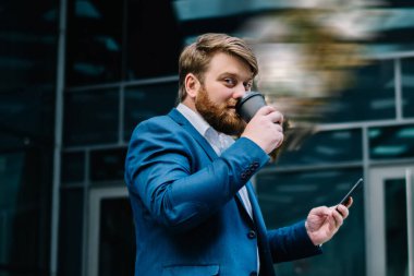 Mavi takım elbiseli, gülümseyen, kahve içen ve elinde akıllı telefonla modern sanatın kurulumunun karşısında duran şişman bir erkek.