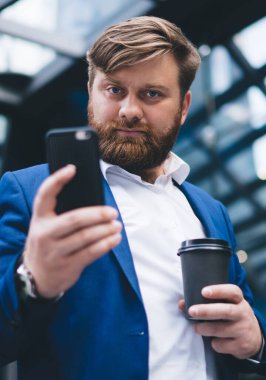 Sakallı, akıllı telefon kullanan, elinde kahve fincanı tutan ve cam çatının altında kameraya bakan kendine güvenen yakışıklı bir yetişkin.