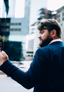 Şık, ciddi iş adamının cep telefonuyla selfie çekerken özgüven dolu bakışları cam iş merkezinin arka planına yarım dönüş yapıyor.