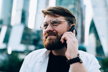Akıllı, entelektüel, gözlüklü, gayri resmi giyinmiş, arka planda modern ayna şehri gökdelenleri olan cep telefonuyla konuşan kızıl sakallı erkek.