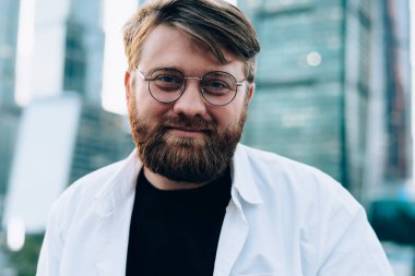 Kırmızı sakallı, beyaz tişörtlü ve siyah tişörtlü, arka planda ayna gökdelenleri olan mutlu, zeki bir erkek.