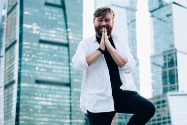 Arka planda şehir merkezindeki gökdelenlerle birlikte ağaç talimi yapan mutlu erkek portresi, dışarıda yoga eğitimi için Vrksasana yapan şık giyimli, pozitif bir adam.
