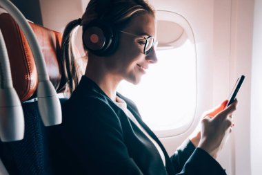 İş sınıfında oturan ve kulaklıklar olumlu ses kitabı dinlerken sohbet için gemide WiFi internet bağlantısı zevk popüler havayolları ile seyahat gülümseyen kadın yan görünümü