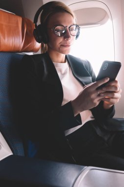 Resmi aşınma giyinmiş kafkas kadın yolcu koltuğuna oturan uçak içinde cep telefonu kullanımı, başarılı kadın okuma kulaklık ile ses kitabı dinlerken cep telefonu ile e-posta aldı