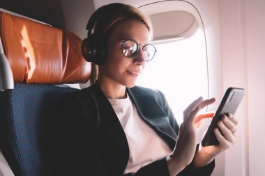 Gemide uçuş sırasında WiFi internete bağlı akıllı telefon ile pencerenin yakınında oturan genç beyaz kadın, App online dükkanda gürültü iptali arama şarkıları için kulaklıkta kadın yolcu