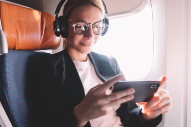 Pencere yakın oturan uçak uçuş sırasında akıllı telefonla okuma haberleri resmi aşınma Kafkas kız, Wi-Fi bağlantısı ve Bluetooth kulaklıklar kullanarak online video izlerken göz giyim iş kadını