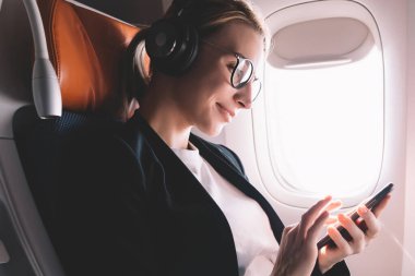 Hipster kız uçuş yolcu cep telefonu uygulamasında müzik şarkıları zevk gürültü iptali için Bluetooth kulaklıklar, gülümseyen kadın uçak kabin içinde kablosuz ağa Smartphone ile bağlı