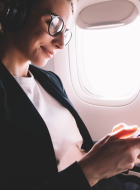 Mutlu gülümseyen hipster kız cep telefonu ile telefon müzik dinleme gemide WiFi bağlı, pozitif kadın yolcu akıllı telefonda iyi kablosuz internet ile ilk sınıfta uçuş zevk