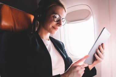 Pozitif genç kadın uçak yolcu koltuğunda oturan ve dijital tablet uygulama ve Bluetooth kulaklıklar üzerinden müzik şarkıları dinleme ile ilginç bir kitap okuma. Gemide kablosuz internet