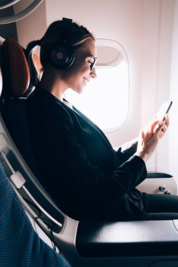 Uçuş sırasında cep telefonu ile dinleme müzik ve tarama ağı zevk uçakta yolcu koltuğuna oturan neşeli kadın müzik sevgilisi yan görünümü. Gemide kablosuz kullanarak genç kadın yolcu