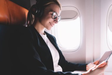 Uçak penceresinde yakın oturan mutlu bir kadın, gemide WiFi internet bağlanırken dijital tablette film izlerken gürültü iptali için gözlük ve kulaklıklar neşeli kadın yolcu