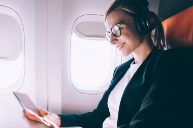 Uçak penceresinde yakın oturan mutlu bir kadın, gemide WiFi internet bağlanırken dijital tablette film izlerken gürültü iptali için gözlük ve kulaklıklar neşeli kadın yolcu