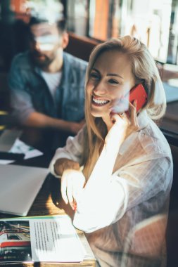 Cafe 'de genç iş arkadaşları ve gazetelerle otururken cep telefonuyla konuşan neşeli genç bir kadının yan görüntüsü.