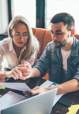 Modern kadın ve erkek takım halinde çalışırken kağıt çizelgelerini tartışıyor ve kafeteryada kağıt ve dizüstü bilgisayarlarla masaya oturuyorlar.