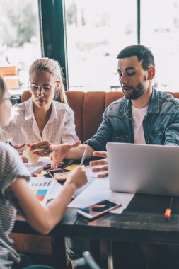 Bir grup modern kadın ve erkek, gündelik giysiler içinde kafeteryada dizüstü bilgisayar ve kağıtlarla oturmuş tartışırken...