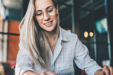 Gözlüklü hoş sarışın, mesajları kontrol ederken masada otururken ve laptopla çalışırken ve kafede kağıtlarla çalışırken.