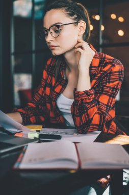 Kafede dizüstü bilgisayar ve not defteriyle otururken ve çalışırken gözlüklü hoş bir kadın kağıt döküman okuyor.