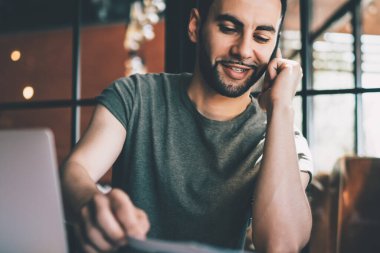 Kafede cep telefonuyla konuşurken dizüstü bilgisayarla masada oturan ve not defterine bakan genç bir adam.