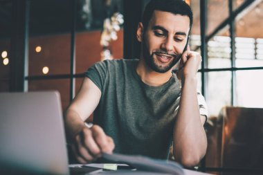 Kafede cep telefonuyla konuşurken dizüstü bilgisayarla masada oturan ve not defterine bakan genç bir adam.