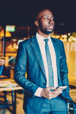 Ofis iç Smartphone ile resmi aşınma ayakta giyinmiş Afrikalı Amerikan işadamı durulması. Düşünceli Dark tenli gurur CEO 'su el cep telefonu ile iş günü planlama