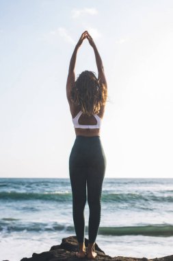 Genç bir kadının arka görünüşü, kurbağa yavrusu pozu verirken vücut bilincini artırıyor. Okyanus kıyısında poz veriyor. Ruh yaratma, sağlıklı yaşam tarzı ve yoga meditasyonuyla uyum kavramının tadını çıkarıyor.