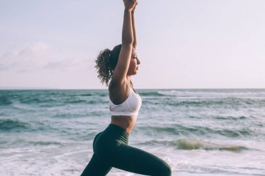 Rastgele eşofman giyen çekici genç bir kadın açık deniz eğitimi sırasında virabhadrasana yapıyor, kadın yoga yapıyor zihinleri rahatlatmak ve ruh uyumunu sağlamak için sağlıklı yaşam tarzından hoşlanıyor.