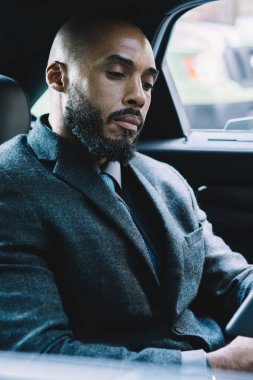 Koyu tenli erkek girişimci resmi giyinmiş. Borsacı web sitesinden finansal dünyadaki yenilikler hakkında e-posta almış. Havaalanına giden yolda bilet kesen adam.