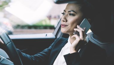 Profesyonel kadın sürücü otomobil yolculuğuna odaklanmış iş görüşmesi saatleri ve akıllı telefon aleti kullanan kadın tüccar hakkında iş arkadaşlarıyla uluslararası telefon görüşmeleri yapıyor.