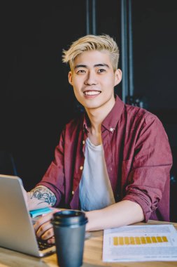 Genç, mutlu Japon hipster adam, sosyal ağlardan, teknoloji ve uzak iş kavramlarından takipçilerle iletişim kurmak için netbook ile modern kampüste otururken gülümsüyor ve başka tarafa bakıyor.