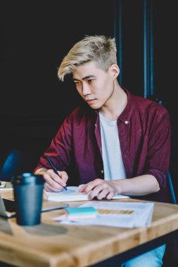 Ciddi Japon hipster adam otomasyon için zaman harcıyor birlikte çalışma alanında bilgi verici webinar izliyor modern dizüstü bilgisayarla ve eğitim için ders kitabına ana notlar yazıyor.