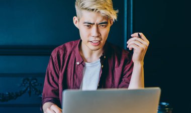 Serbest çalışan Çinli bir gencin modern dizüstü bilgisayardaki yazılımla sorunu olduğunu merak ettim. Kablosuz internet bağlantısı kullanarak internetten Webinar izleyen kafası karışmış bir hipster.