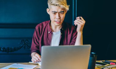 Serbest çalışan Çinli bir gencin modern dizüstü bilgisayardaki yazılımla sorunu olduğunu merak ettim. Kablosuz internet bağlantısı kullanarak internetten Webinar izleyen kafası karışmış bir hipster.