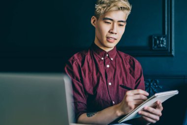 Elinde not defteri tutan ve dizüstü bilgisayarında oturan düşünceli sarışın Japon hipster çocuk. Netbook 'ta çalışırken not defterine not alan Çinli öğrenci.