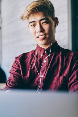 Neşeli Asyalı öğrenci kulaklıklarıyla modern bilgisayarda komik dersler seyrediyor. Modern mekanda oturan Çinli genç adam dijital bilgisayarda kablosuz internet kullanarak serbest çalışıyor.