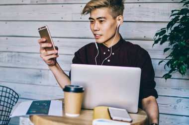 Havalı hipster adam akıllı telefon uygulaması aracılığıyla mesaj aldı. Elektronik kulaklıklar aracılığıyla müzik listesi dinlerken kafeteryada modern dizüstü bilgisayarla oturuyordu.