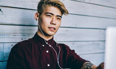 Düşünceli Japon hippi öğrenci kulaklıkla bilgisayarda Webinar 'ı seyrediyor modern birlikte çalışma alanında oturuyor. Konsantre Çinli genç adam internet üzerinden internet üzerinden bilet alıyor.