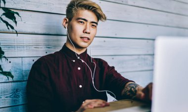 Düşünceli Japon hippi öğrenci kulaklıkla bilgisayarda Webinar 'ı seyrediyor modern birlikte çalışma alanında oturuyor. Konsantre Çinli genç adam internet üzerinden internet üzerinden bilet alıyor.