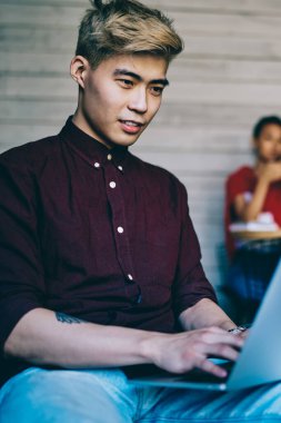 Neşeli Çinli hippi öğrenci modern dizüstü bilgisayarda ders çalışması için günlük giyim metni yazıyor ve birlikte çalışma alanındaki kablosuz internet bağlantısını kullanarak internet sitelerinde arama yapıyor.