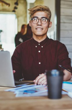 Görme düzeltmesi için optik gözlüklü Çinli hippi öğrenci ahşap masaüstünde dijital bilgisayarla otururken web sitesinde yayınlanması için yaratıcı fikirler düşünüyor.