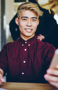 Modern uzayda kameraya bakarken akıllı telefonu elinde tutan neşeli Çinli hippi blogcu portresi. Sarı saçlı genç adam cep telefonuna SMS mesajı gönderiyor.