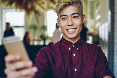 Neşeli gülümseyen Asyalı öğrenci elinde cep telefonuyla masaüstünde oturuyor ve 4G internet bağlantısı kullanıyor, mutlu Japon hipster adam cep telefonuyla çevrimiçi hizmetten geri bildirim bekliyor.