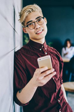 Modern cep telefonunu elinde tutan ve boş zamanlarında kameraya gülümseyen optik gözlük takan binlerce yıllık erkek blog yazarının yarım boy portresi.