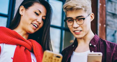 Neşeli hipster Asyalı öğrenciler ellerinde modern akıllı telefonlar ve çevrimiçi video izliyorlar şehir ortamında dikiliyorlar. Pozitif Japon arkadaşlar hücresel ağlarda tasarım profillerini tartışıyorlar.